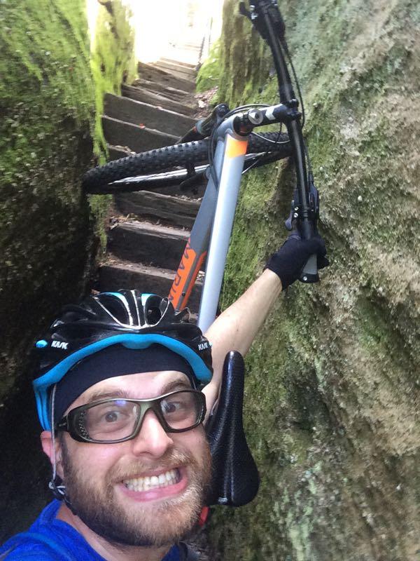 A person in cycling gear smiling while holding a mountain bike, standing between two rocky walls that have moss on them, with wooden steps visible in the background leading upward. The scene appears to be in a narrow, natural passageway. Black Mountain mountain bike trail.