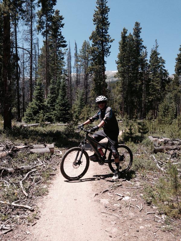 A person wearing a helmet and cycling gear is standing on a dirt trail surrounded by trees in a forest setting. They are holding onto a mountain bike and facing the camera, with patches of sunlight filtering through the trees. The background features a mix of tall pine trees and scattered logs on the ground. Peaks Trail mountain bike trail.