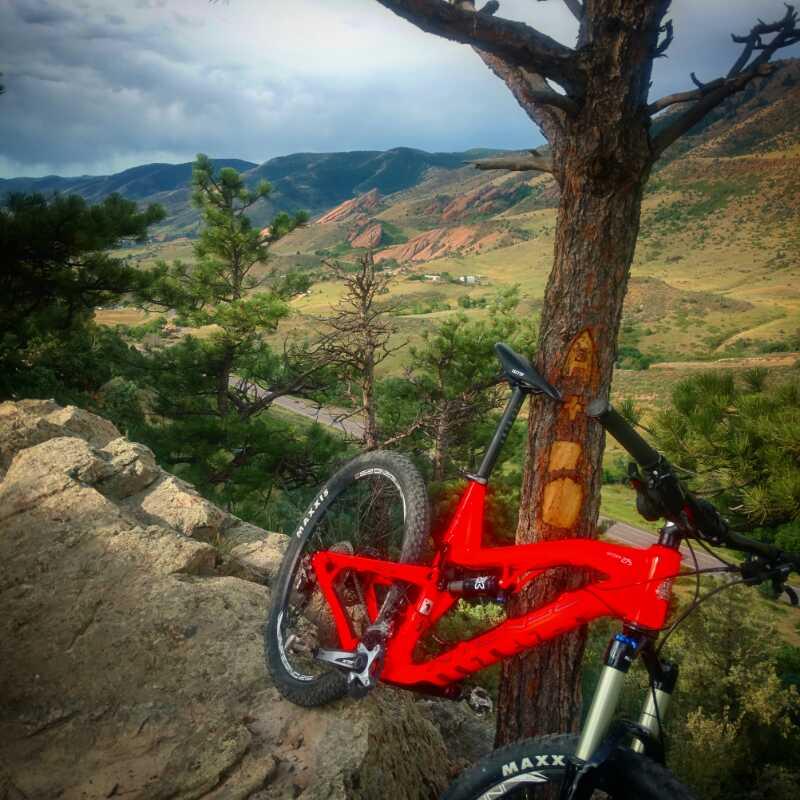 A vibrant red mountain bike leaned against a tree on a rocky outcrop, overlooking a scenic valley dotted with green trees and sloping hills. The sky is partly cloudy, adding depth to the landscape, while the foreground features rugged rocks and the bike's tire prominently displaying the brand. Red Rocks / Dakota Ridge mountain bike trail.