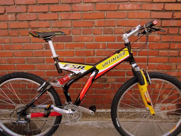 Specialized FSR: A mountain bike featuring a sleek black and yellow frame with the brand name "Specialized" prominently displayed. The bike has a suspension system, thick tires for rough terrain, and a vibrant red and black seat. It is positioned against a rustic red brick wall.
