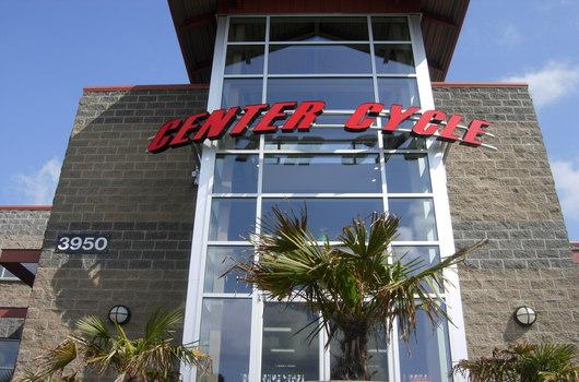 Exterior view of a storefront for "Center Cycle," featuring large glass windows and a prominent sign displaying the business name in red. The building includes palm trees in the foreground and the address number 3950 near the entrance. Clear blue skies are visible above.