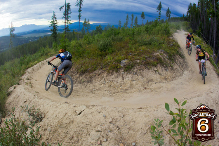 A group of mountain bikers riding on a dirt trail surrounded by lush greenery and trees, with a scenic view of mountains and a valley in the background. The scene is set under a cloudy sky, conveying an adventurous atmosphere.
