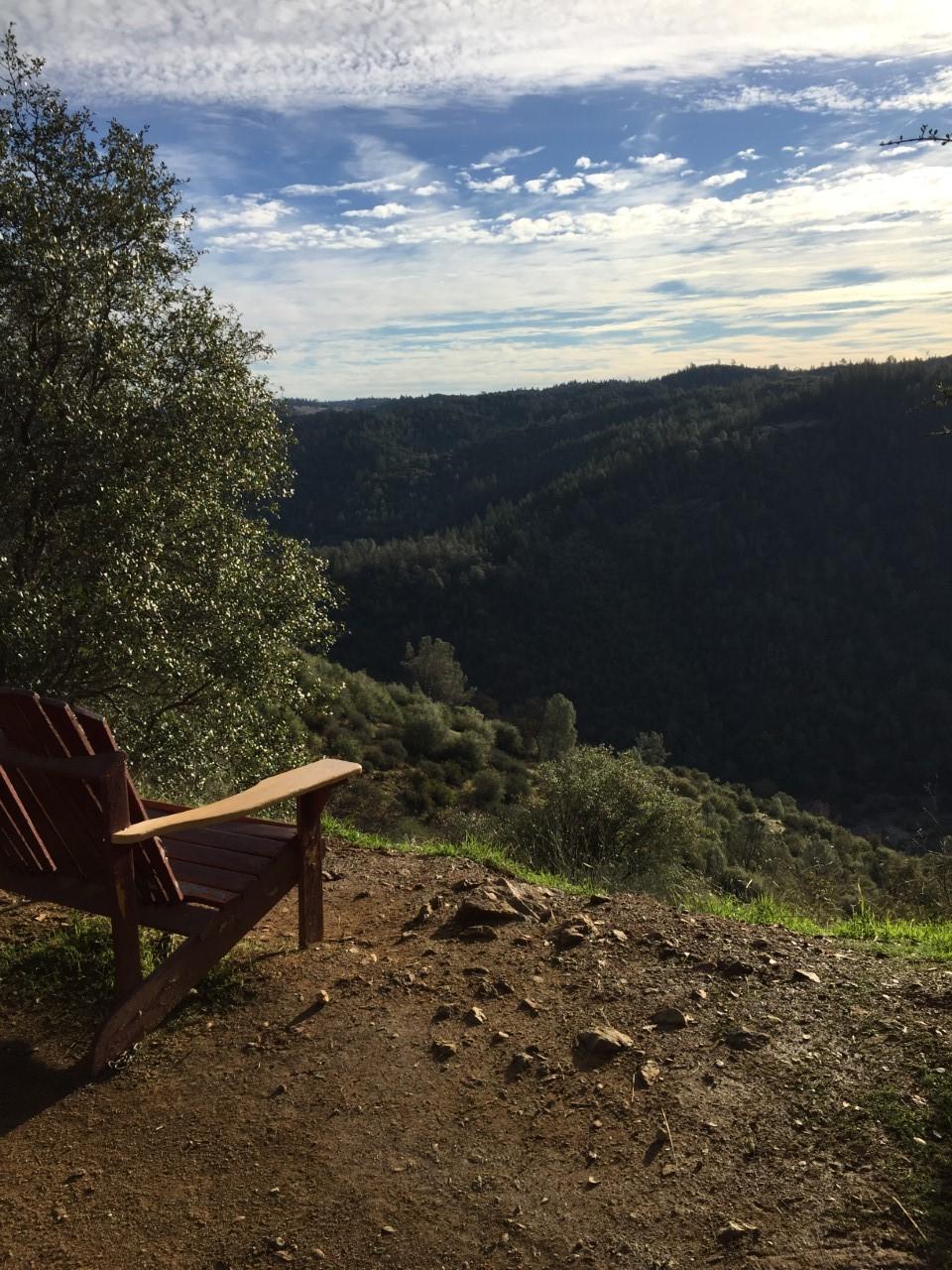 A wooden Adirondack chair sits on a dirt path overlooking a lush green valley and rolling hills under a partly cloudy sky. The scene conveys tranquility and natural beauty, inviting relaxation in a serene outdoor setting. Stagecoach / Flood / Manzanita mountain bike trail.