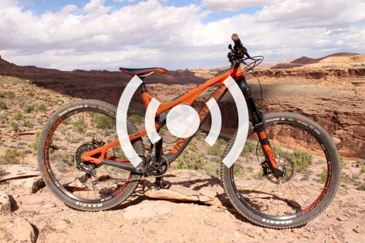 An orange mountain bike is positioned on rocky terrain, with a scenic backdrop of canyons and hills under a partly cloudy sky. The bike's tires and frame are prominently displayed, highlighting its rugged design suitable for outdoor adventures.