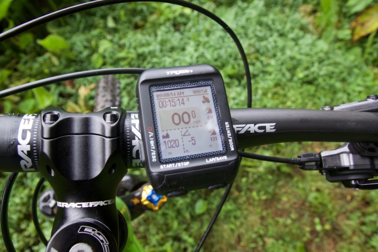 A close-up view of a mountain bike's handlebars, featuring a digital display with data such as time, speed, and elevation, set against a backdrop of green foliage. The handlebars include branded components, and the display shows various metrics related to a ride.