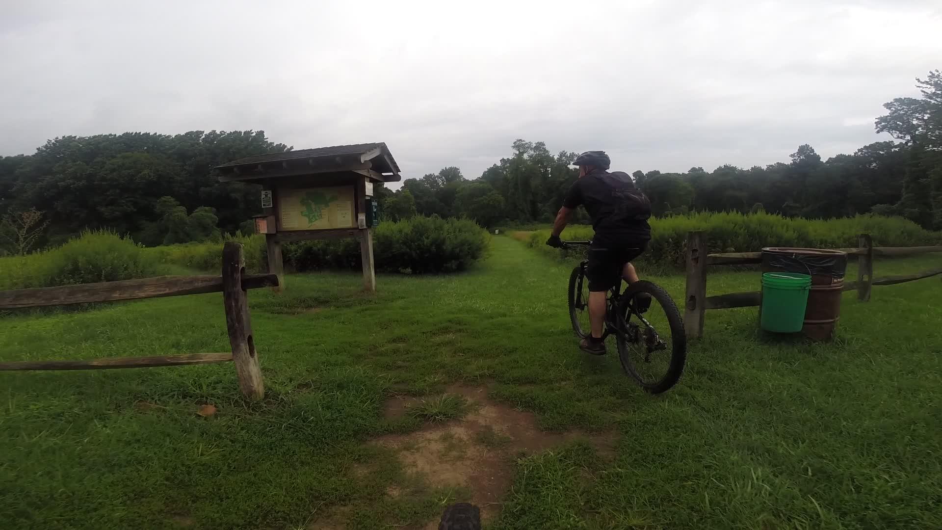A mountain biker riding towards a trailhead sign in a lush, green outdoor area, with a wooden fence and trash barrels nearby under a cloudy sky. Huber Woods mountain bike trail.