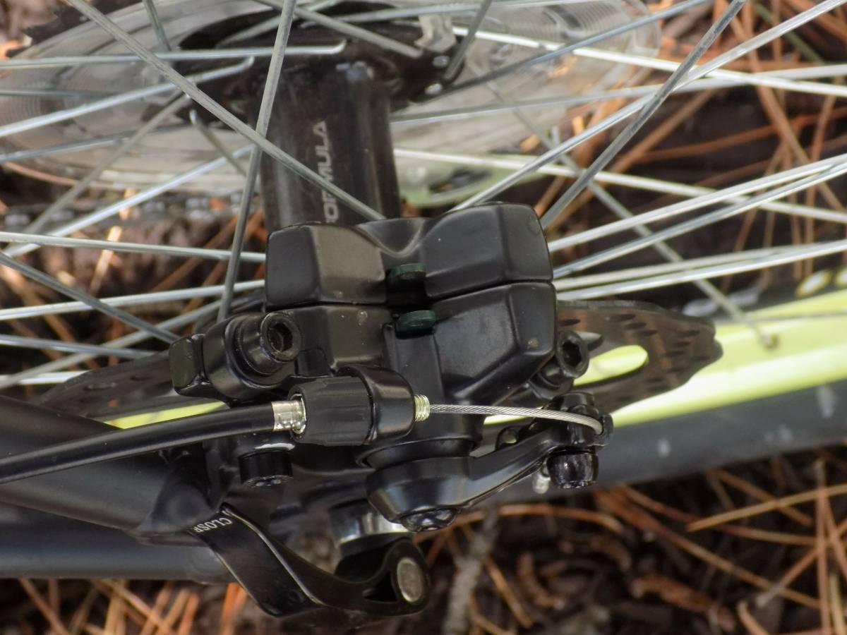Motobecane Fantom29 Sport: Close-up view of a bicycle disc brake assembly, showing the brake caliper, rotor, and nearby wheel spokes. The image captures the details of the brake components against a backdrop of pine needles.