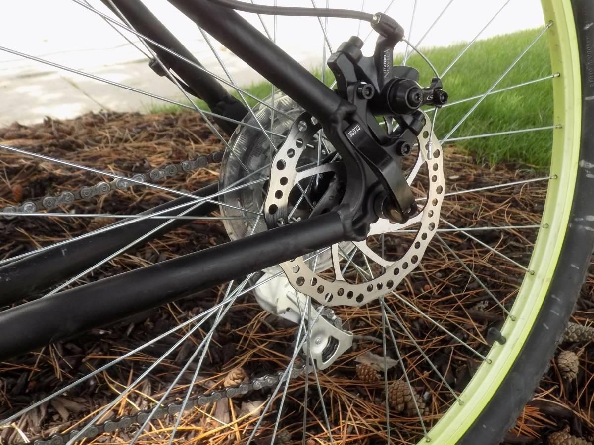 Motobecane Fantom29 Sport: Close-up view of a bicycle's rear wheel hub, featuring a disc brake system. The image shows the metal brake caliper, the brake disc, and the bike chain, with pine needles and grass in the background.