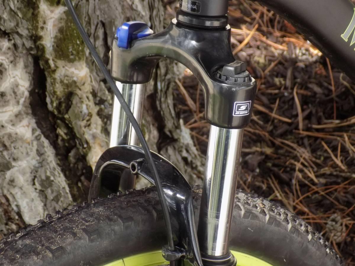 Motobecane Fantom29 Sport: Close-up of a mountain bike front suspension fork and wheel, with a textured tire and a background of a tree trunk and forest floor debris. The fork features metallic silver stanchions, a black casing, and an adjustable knob at the top.
