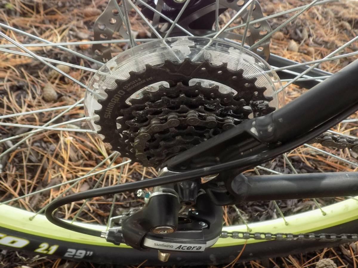 Motobecane Fantom29 Sport: Close-up view of a bicycle's rear gear system, showcasing the Shimano Acera derailleur and cassette. The background features pine needles and earthy textures, indicating an outdoor setting.