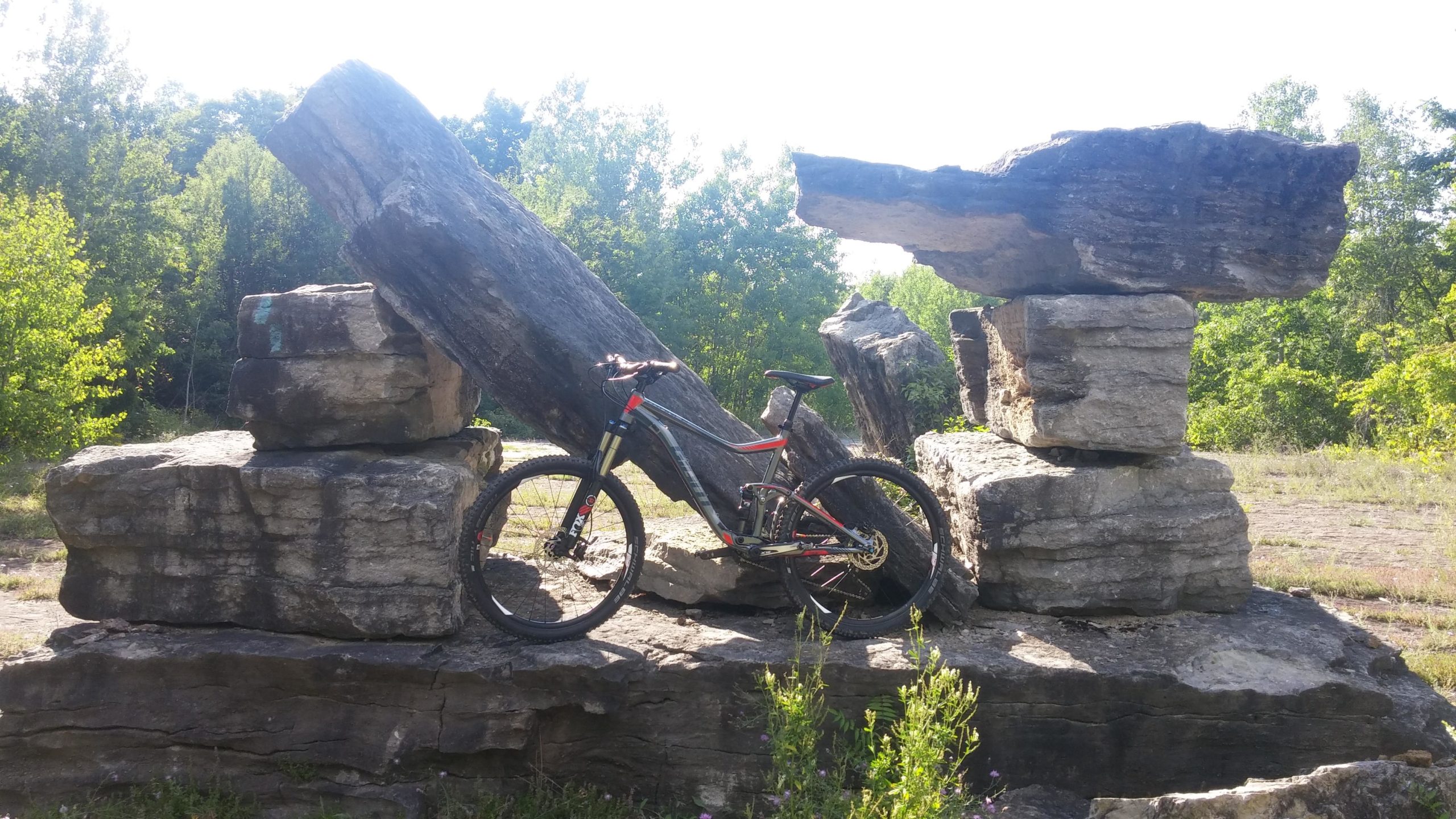 A mountain bike leaned against a large rock formation in a natural outdoor setting, surrounded by greenery and trees in the background. Thatcher State Park mountain bike trail.