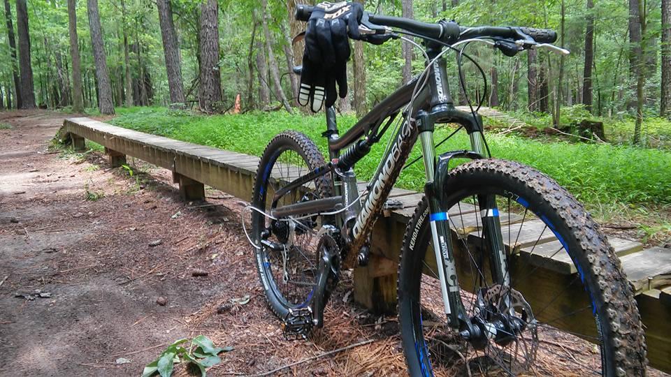 Diamondback Recoil: A mountain bike resting on a wooden trail in a lush green forest, with trees in the background and a patch of grass beside the path.