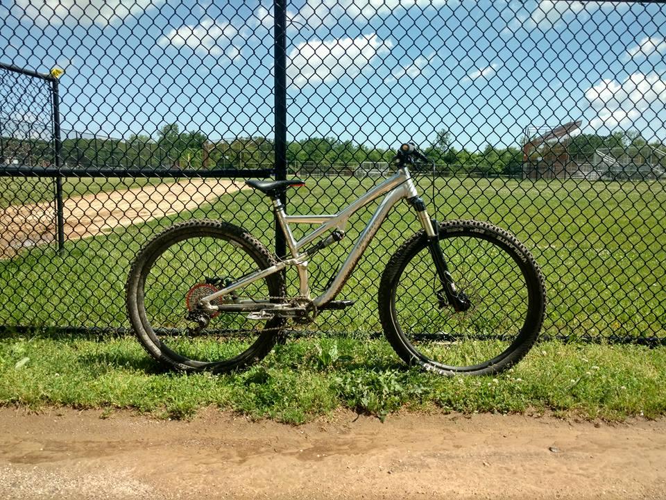 Specialized Stumpjumper FSR Expert: A silver mountain bike leaning against a black chain-link fence, with a baseball field in the background under a clear blue sky. Grass and dirt are visible in the foreground.