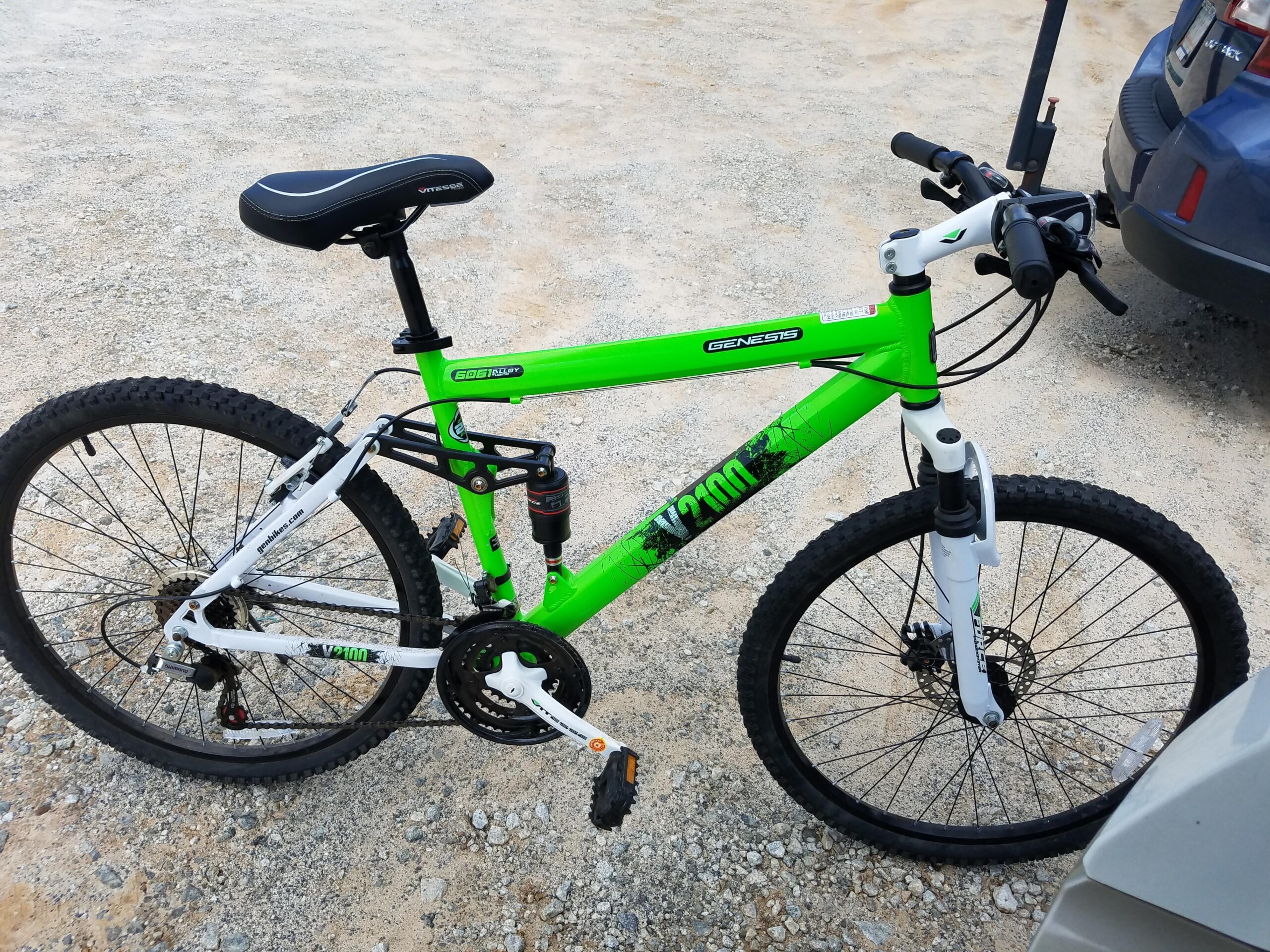 Gensis: A bright green and white mountain bike with black tires and a comfortable seat, parked on a gravel surface. The bike features a sturdy frame, suspension, and disk brakes. There is a blue car partially visible in the background.