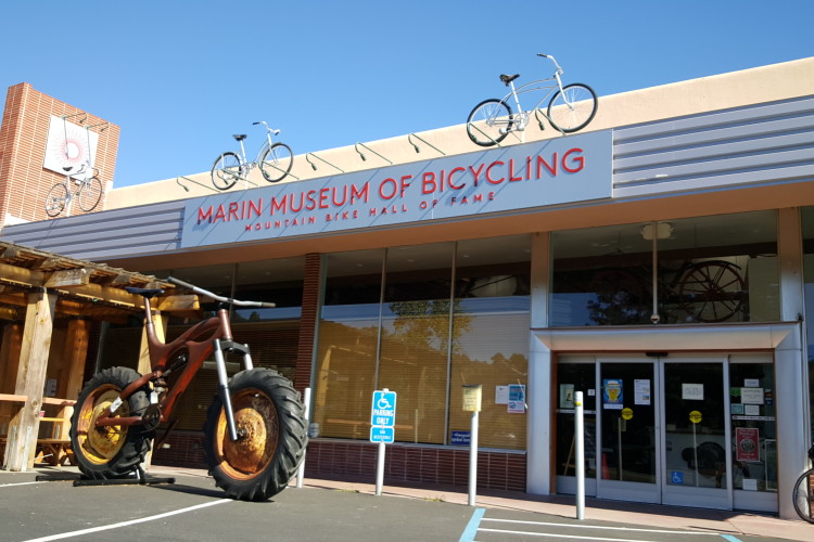 Exterior view of the Marin Museum of Bicycling, featuring a large decorative bicycle sculpture in front and bicycles displayed on the roof. The building has a modern design with large windows and a prominent sign reading "Marin Museum of Bicycling - Mountain Bike Hall of Fame." The surrounding area includes outdoor seating and accessible parking.