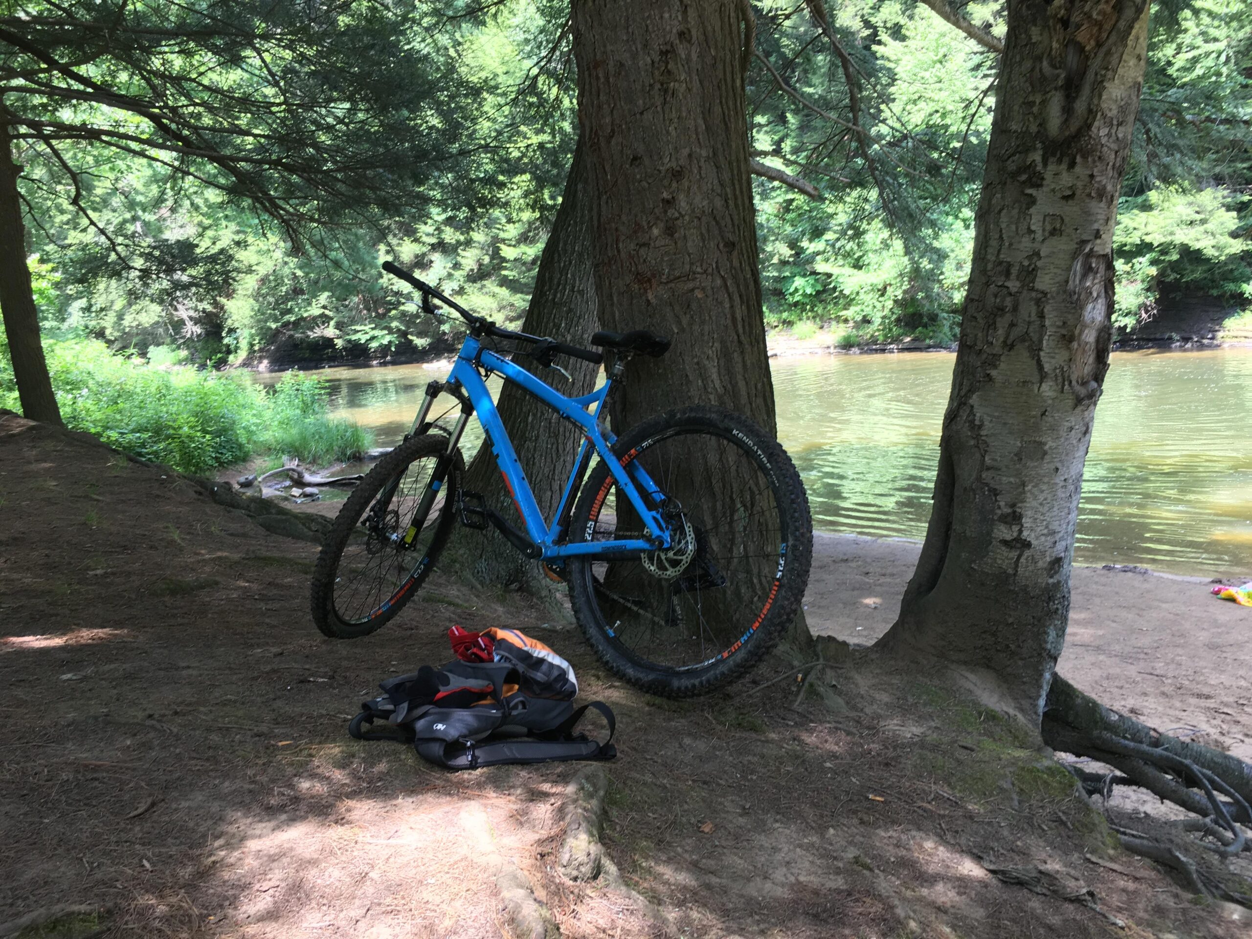Diamondback Hook: A blue mountain bike leaning against a tree near a riverbank, with lush greenery in the background and a small sandy area visible by the water. A backpack is resting on the ground nearby.