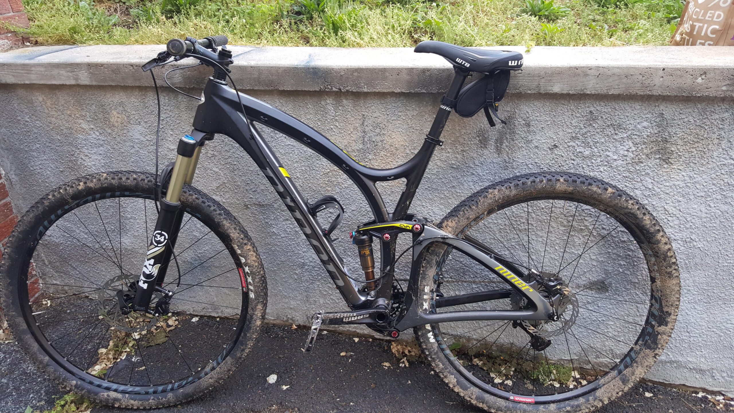 Niner Jet 9 RDO: Mountain bike leaning against a textured gray wall, featuring a lightweight frame, thick tires with mud, and front suspension. The bike has visible components like disc brakes and a saddle bag. Green grass and a hint of greenery are visible in the background.