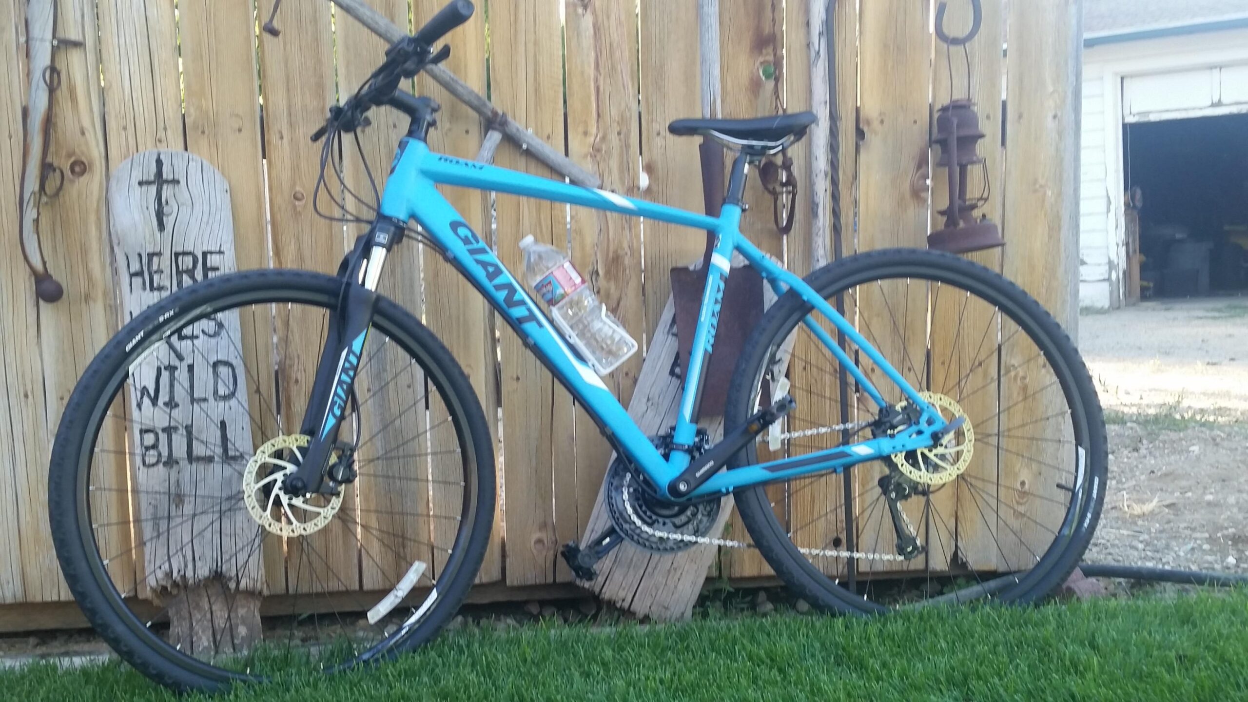 Giant Roam 0: A blue Giant mountain bike leaning against a wooden fence, with a bottle of water secured in the frame. The backdrop features a wooden sign that reads "HERE LIES WILD BILL," along with a hanging lantern and rustic decor. The scene includes green grass in the foreground and a glimpse of a garage in the background.