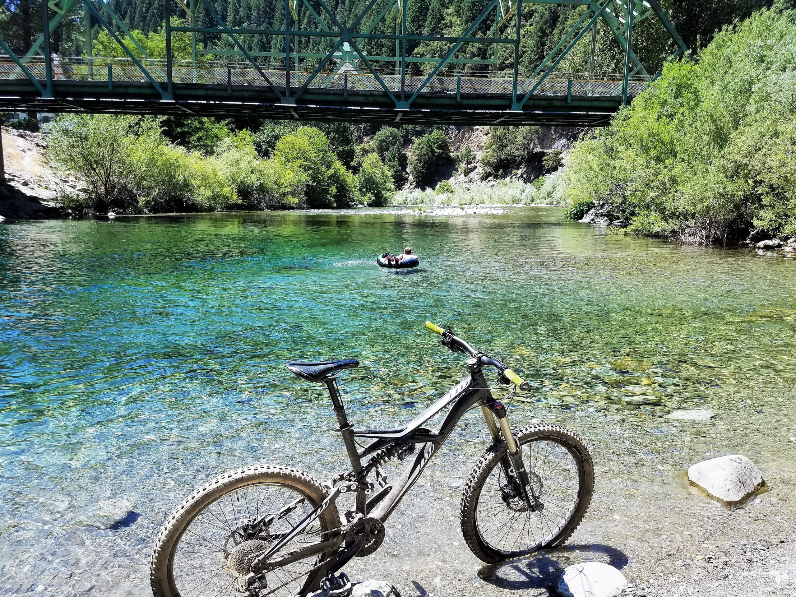 Specialized Enduro Evo: A mountain bike parked on the shore of a clear, calm river, with a green metal bridge spanning above. In the background, a person in a flotation device is seen floating in the water, surrounded by lush greenery and hills. Sunlight glistens on the water's surface, creating a tranquil outdoor scene.