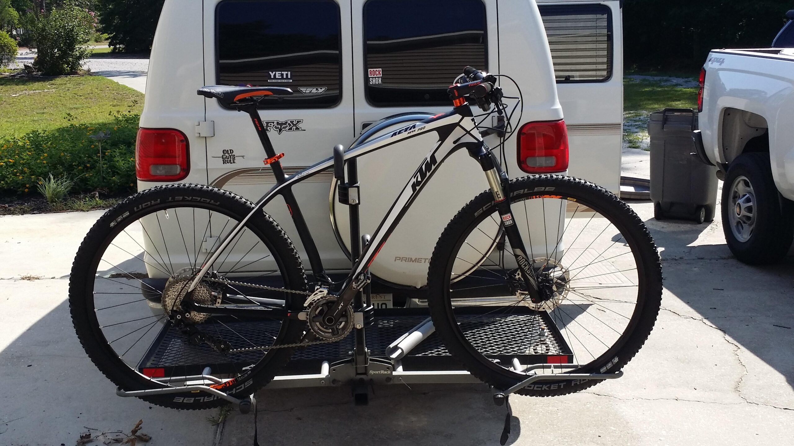 Ktm Aera Pro: A mountain bike secured to a hitch-mounted bike rack on the back of a vehicle. The bike features a sleek design with black and orange accents, large tires, and a visible drivetrain. In the background, a white van is parked, along with a truck and some greenery. The scene is set in a driveway with a sunlit atmosphere.