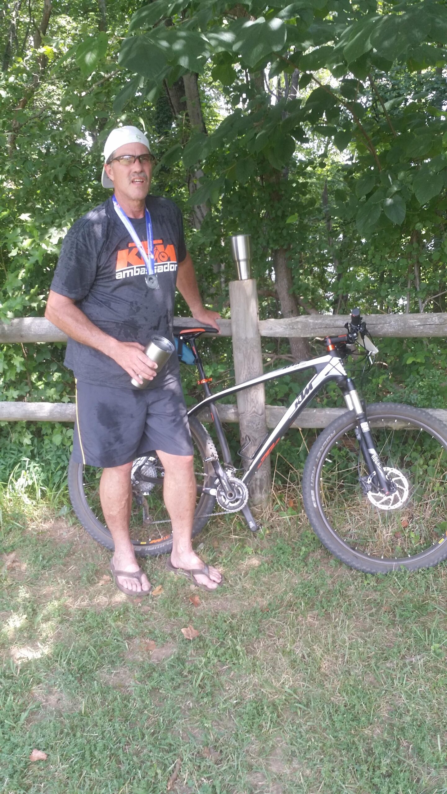 Ktm Aera Pro: A man wearing glasses, a sports t-shirt with "KTM Ambassador" printed on it, and shorts stands next to a mountain bike. He has a medal hanging around his neck and is holding a drink in a stainless steel tumbler. The background features lush greenery and a wooden fence. The man appears to have just finished a cycling activity, as he is visibly sweating.