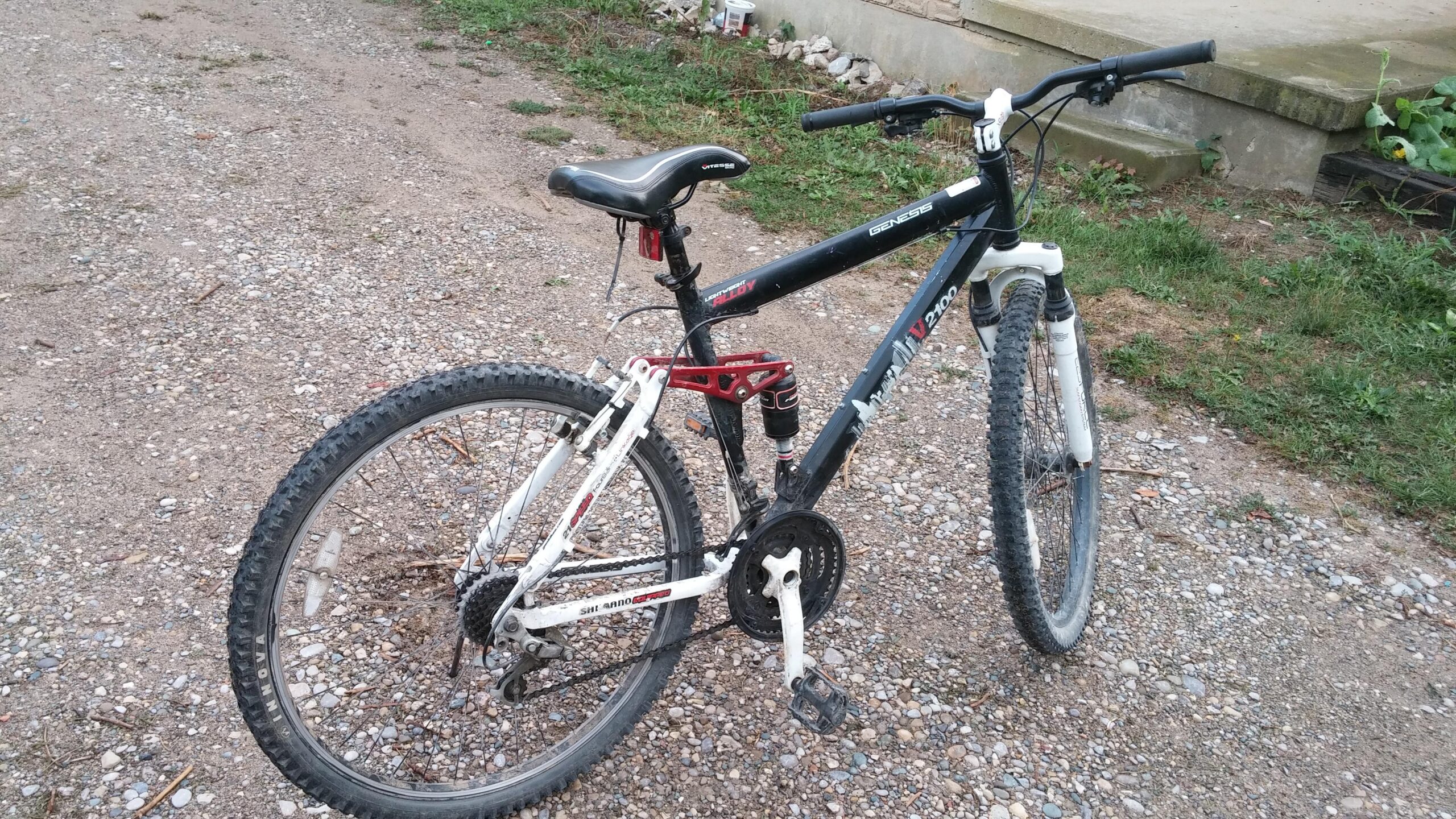 Genesis V2100: Alt text: A black and white mountain bike parked on a gravel path, with a partially grassy area and a building in the background. The bike features a dual suspension frame and visible gear components.