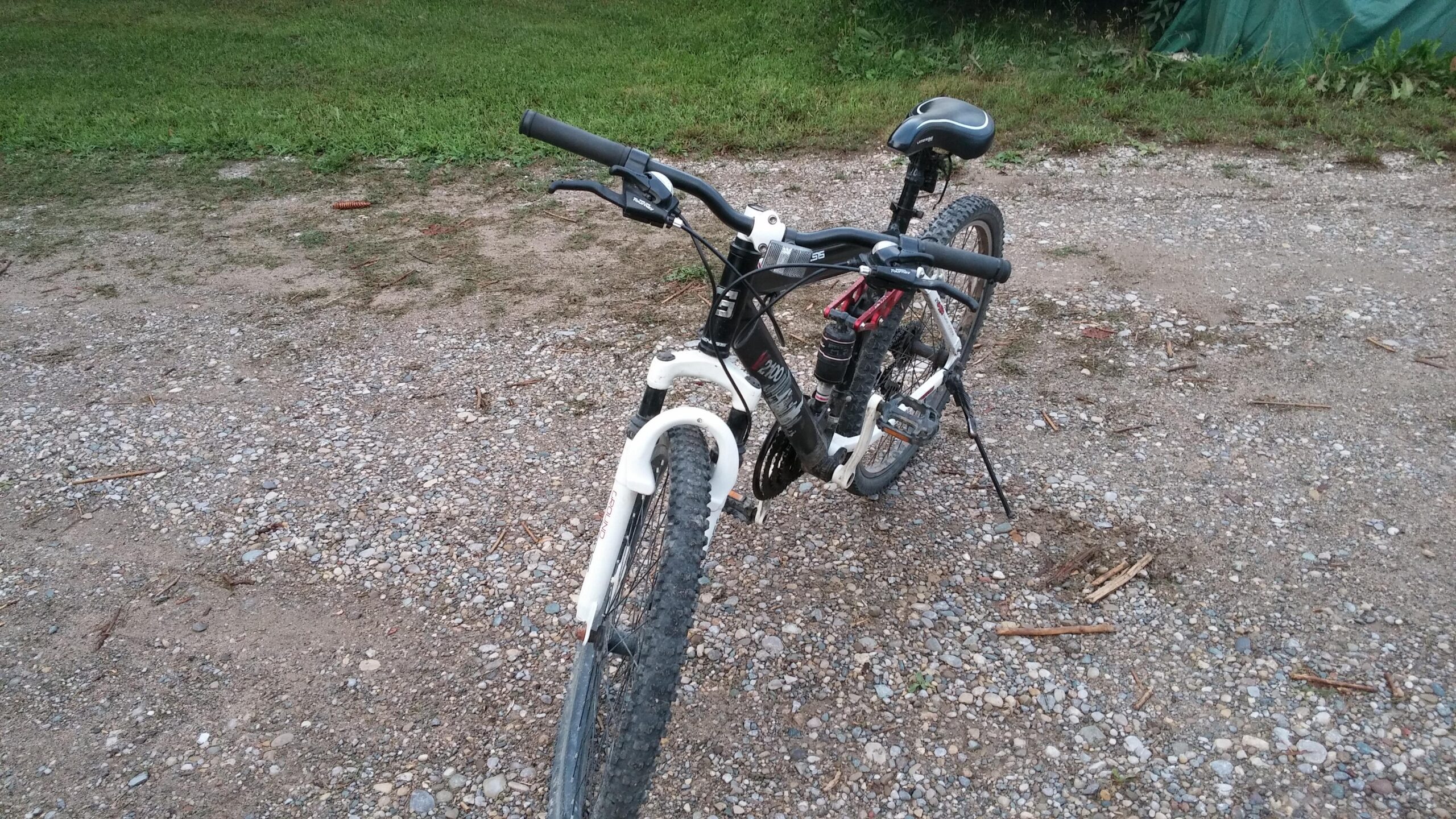 Genesis V2100: A close-up view of a mountain bike resting on a gravel surface. The bike features a black and white frame, with thick, knobby tires designed for off-road terrain. The handlebars are fitted with grips, and there's a small water bottle attached to the frame. In the background, grass and a patch of dirt are visible.