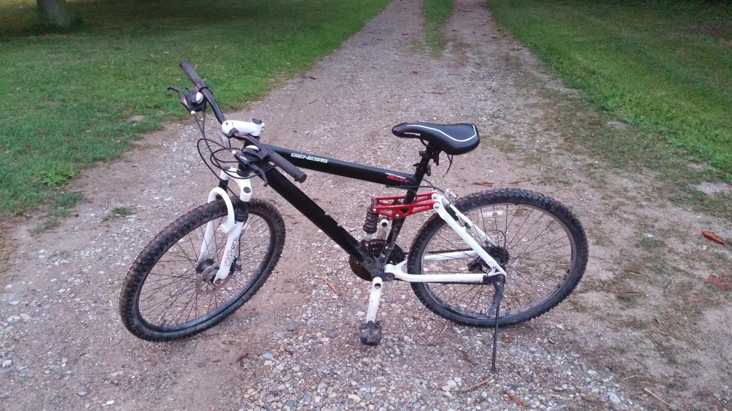 Genesis V2100: A black and white mountain bike parked on a gravel path, surrounded by green grass. The bike features a red rear suspension, thick tires, and a comfortable black seat. 