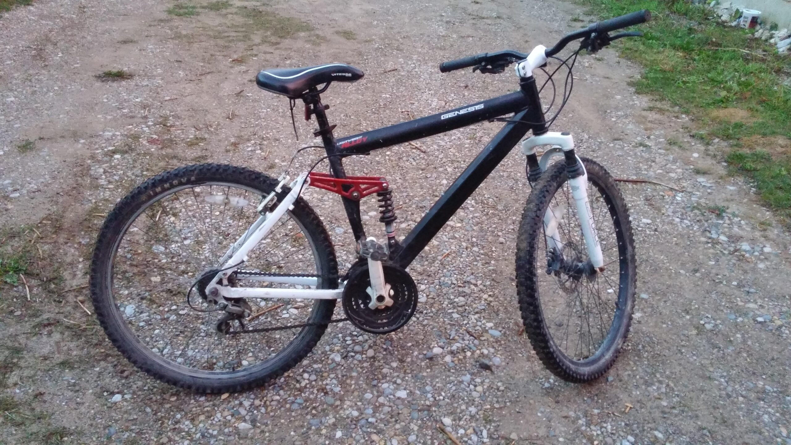 Genesis V2100: A black and white mountain bike parked on a gravel path, featuring a front suspension and a visible rear shock absorber. The bike has thick, knobby tires suitable for off-road riding and is angled to show its frame and components. The background includes patches of grass and a slightly blurred area indicating more gravel.