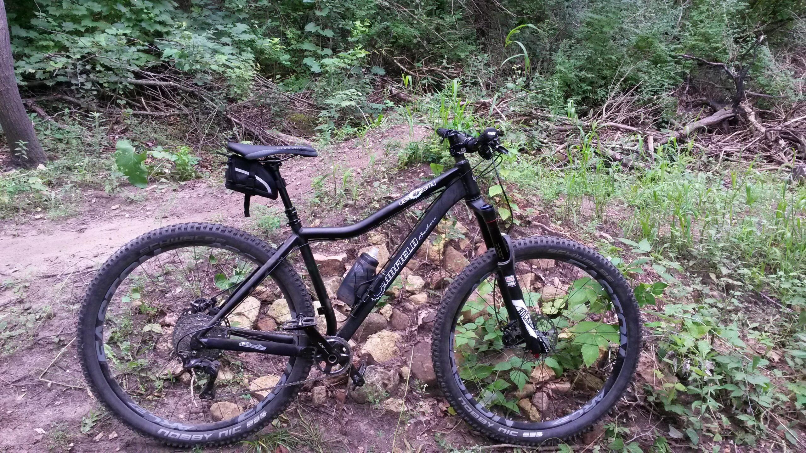 Canfield Brothers Yelli Screamy: A black mountain bike parked on a rocky trail surrounded by lush greenery and trees, with a small black pouch attached to the seat.