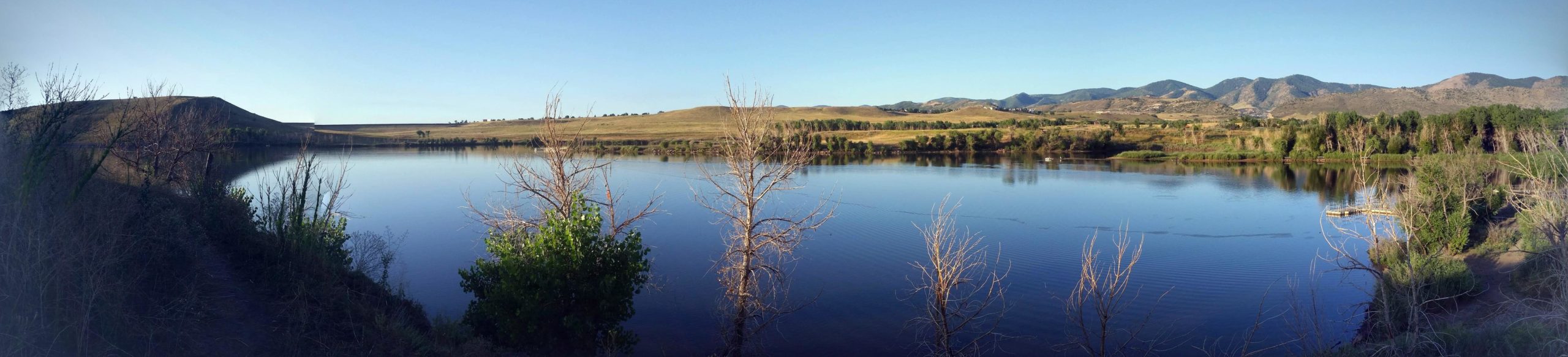 A panoramic view of a tranquil lake surrounded by lush greenery and rolling hills under a clear blue sky. The water reflects the landscape, creating a serene atmosphere, with bare trees lining the shore and distant mountains in the background. Bear Creek Lake Park mountain bike trail.