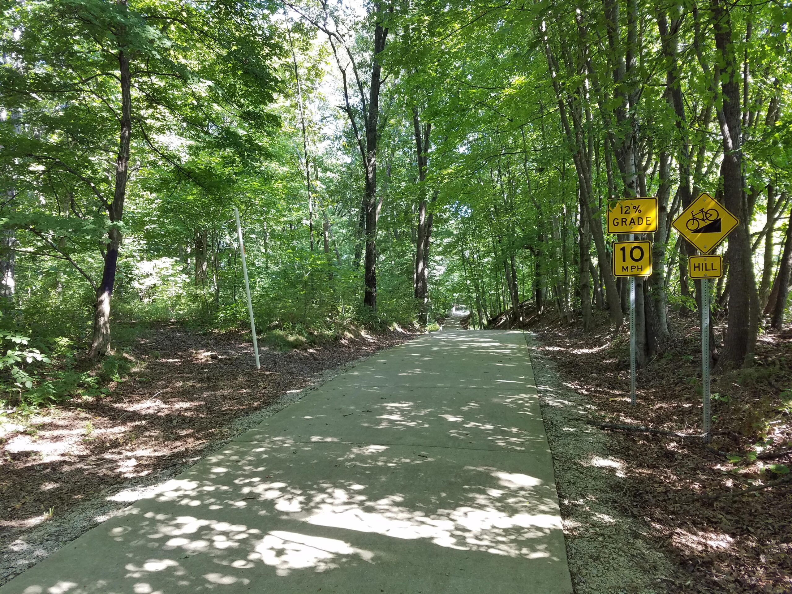 A paved trail surrounded by lush green trees, with signs indicating a 12% grade and a speed limit of 10 mph for cyclists, along with a warning for a hill ahead. Sunlight filters through the canopy, creating dappled shadows on the path. Calico Multi-Use Trail mountain bike trail.