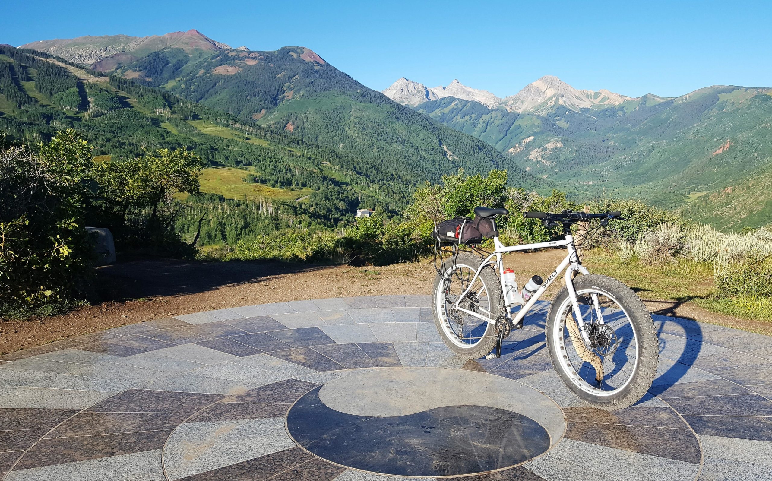 A mountain bike parked on a scenic overlook surrounded by lush greenery and mountain views under a clear blue sky. The ground features a patterned stone design with a circular element in the center. Rim Trail mountain bike trail.