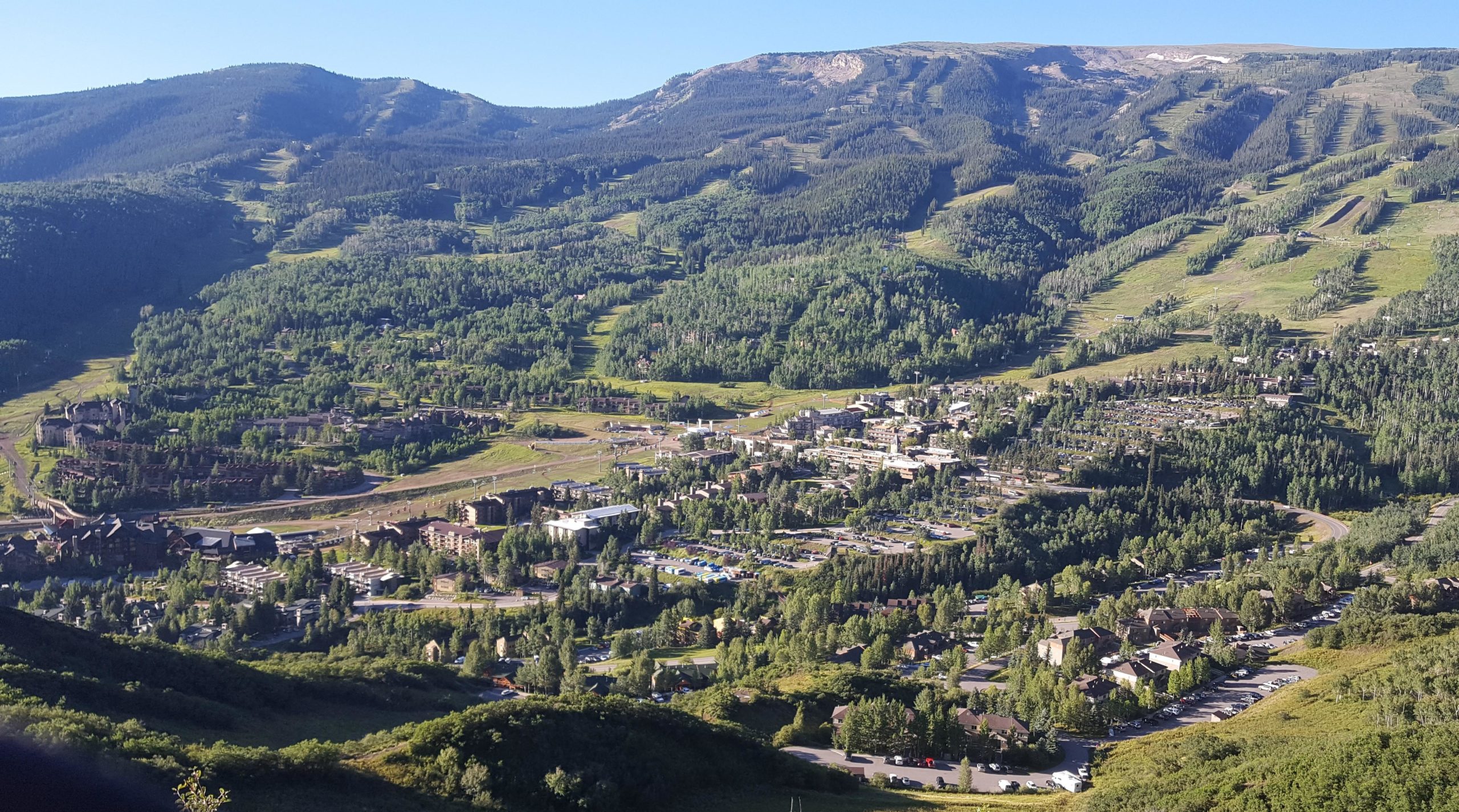 A panoramic view of a mountainous landscape featuring lush green hills and a small town nestled in the valley below. The image showcases vibrant trees and residential buildings, with ski trails visible on the slopes in the background. The sky is clear and blue, suggesting a sunny day. Rim Trail mountain bike trail.