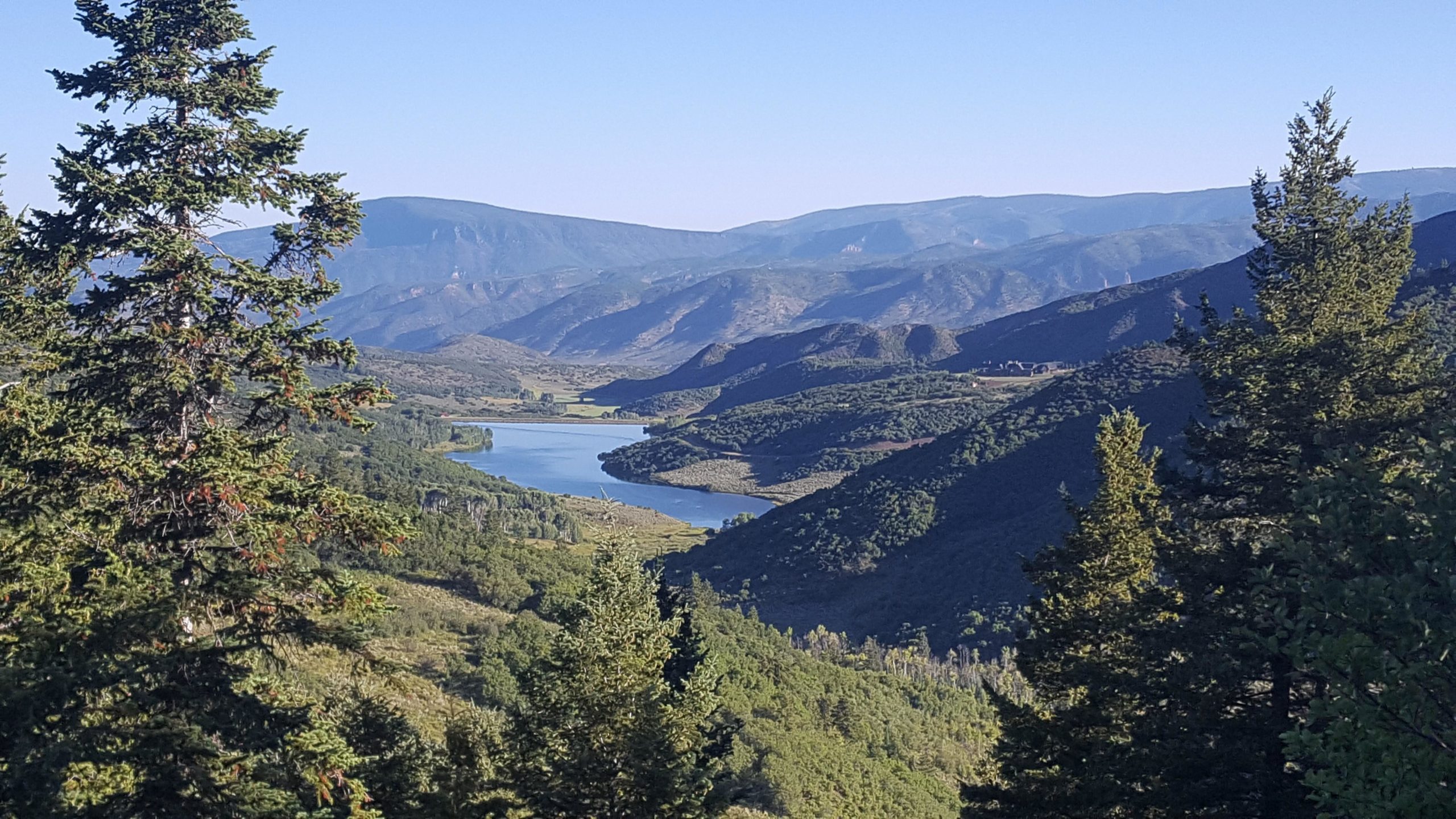 A panoramic view of a serene landscape featuring a winding river surrounded by lush green hills and mountains under a clear blue sky. Pine trees are visible in the foreground, framing the picturesque scene. Rim Trail mountain bike trail.