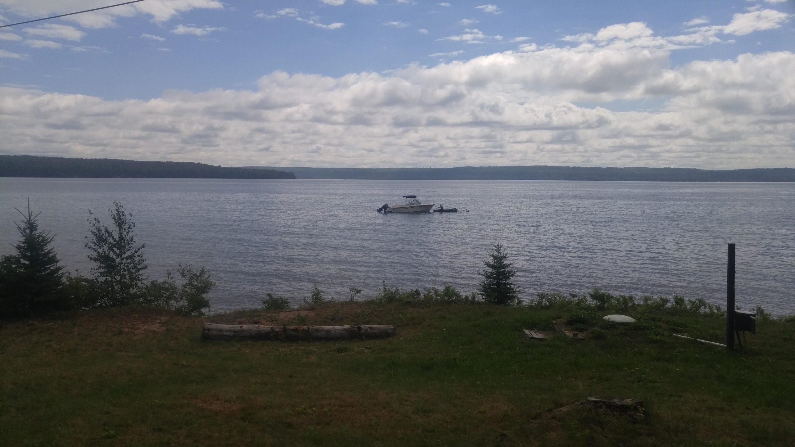 A scenic view of a calm lake with a single boat floating on the water, surrounded by trees and grassy shoreline under a partly cloudy sky. Grand Island mountain bike trail.