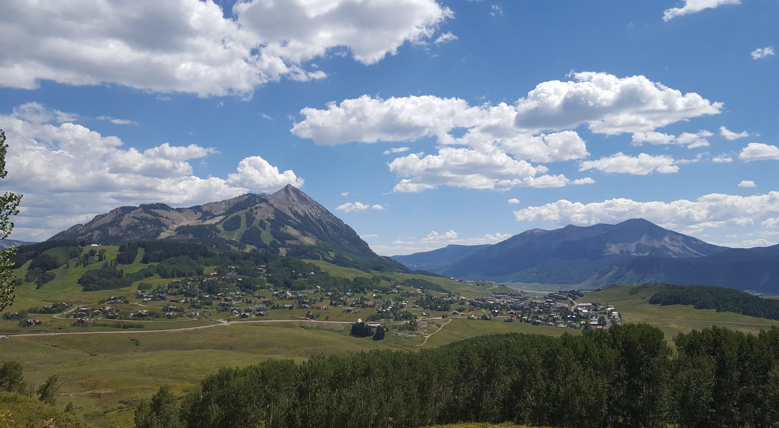 A picturesque mountain landscape featuring a valley dotted with houses, surrounded by lush greenery and rolling hills. Majestic peaks rise in the background under a bright blue sky with fluffy white clouds, creating a serene and scenic view. Snodgrass mountain bike trail.