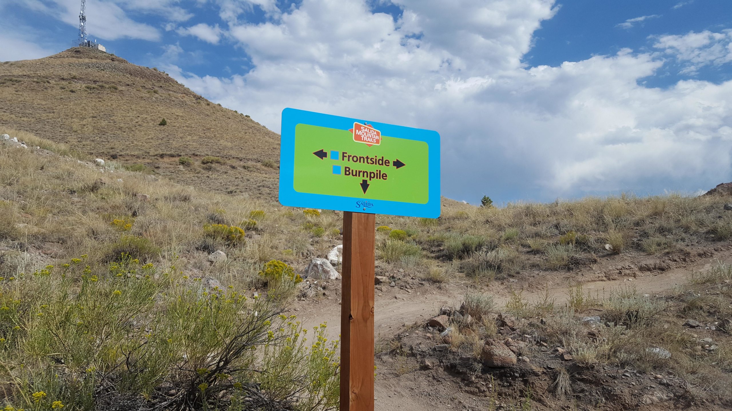 A sign along a hiking trail indicating directions to "Frontside" and "Burnpile," surrounded by dry grass and rocky terrain, with a telecommunications tower on a hill in the background under a partly cloudy sky. Arkansas Hills mountain bike trail.
