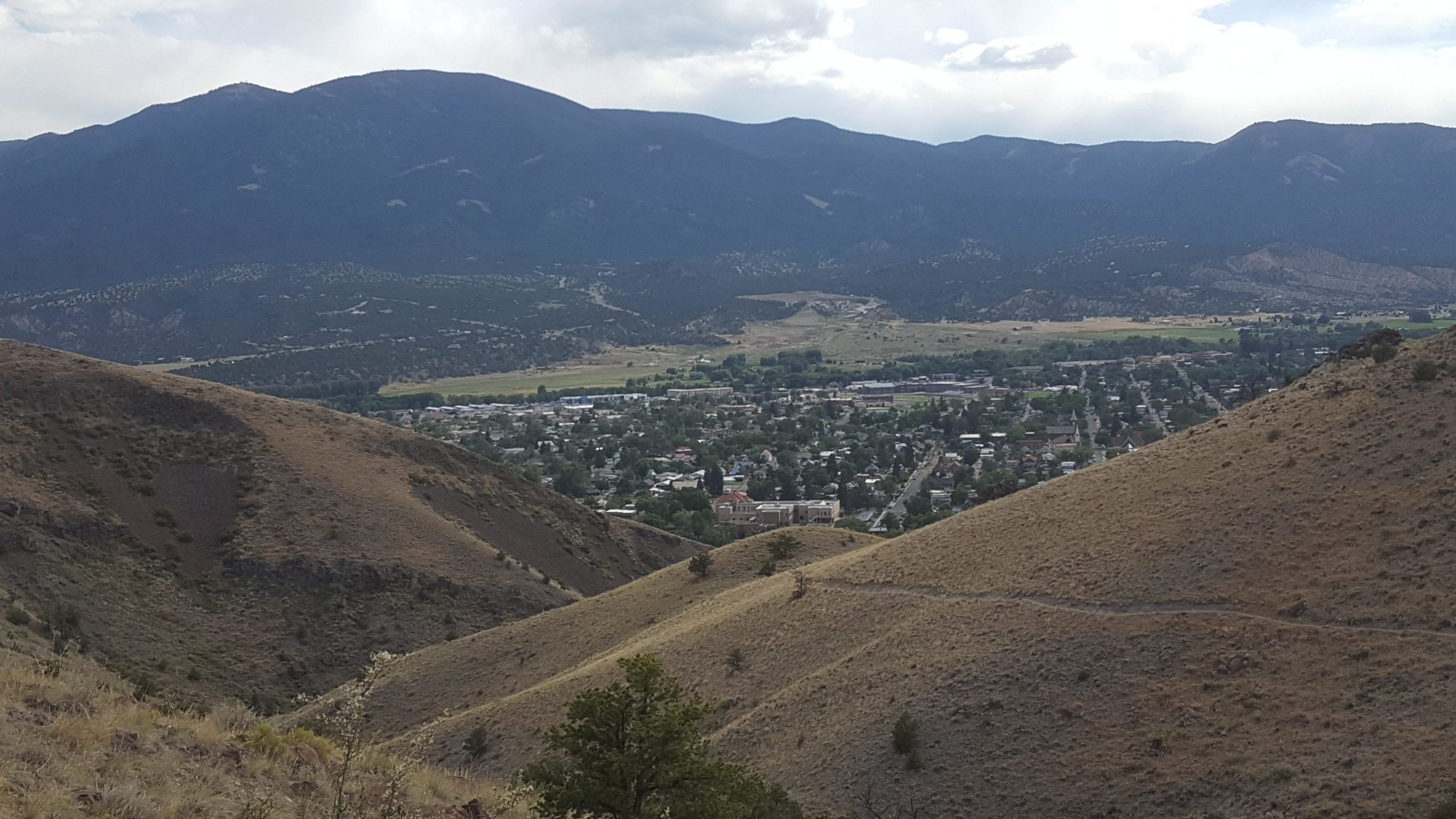 A panoramic view of a small town nestled in a valley, surrounded by rolling hills and mountains under a cloudy sky. The foreground features grassy terrain and shrubs, while the background showcases distant mountain ranges and patches of greenery in the valley below. Arkansas Hills mountain bike trail.