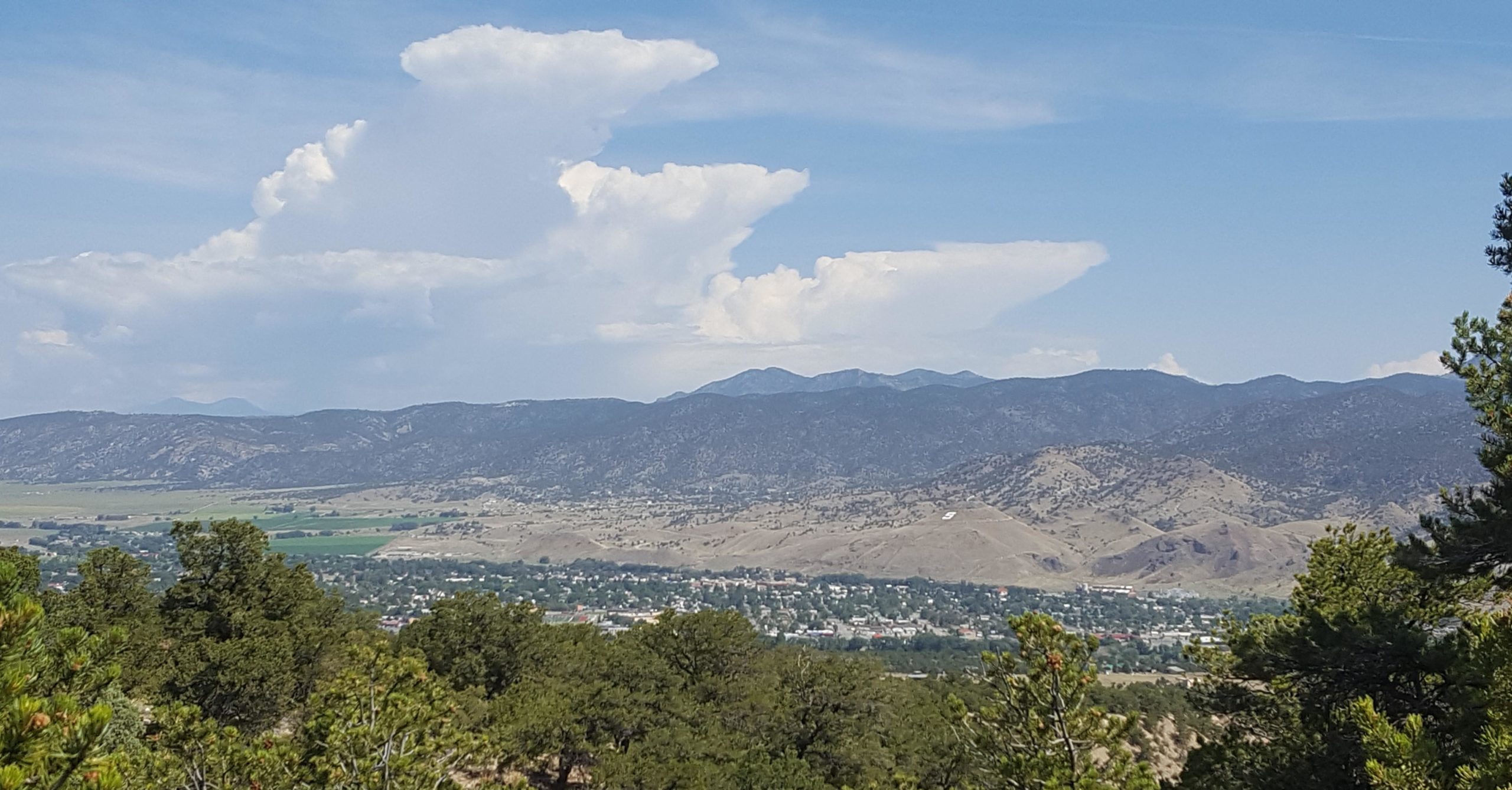 A panoramic view of rolling hills and mountains under a partly cloudy sky. In the foreground, green trees frame the view, while a valley below shows a small town and agricultural fields. The distant mountains create a scenic backdrop against the blue sky. Methodist Mountain mountain bike trail.