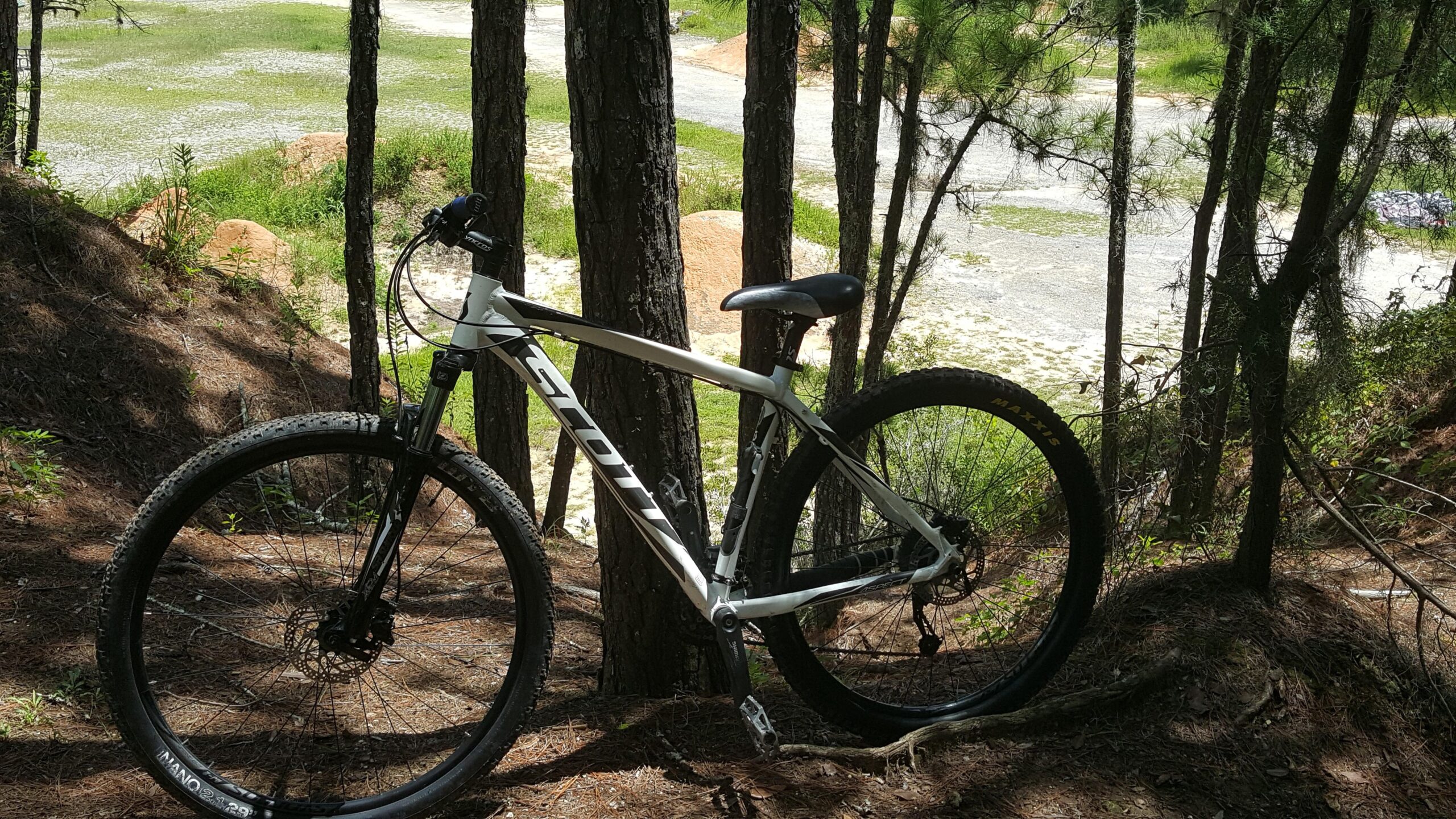 Scott aspect 940: A mountain bike leaning against a tree in a wooded area, with a view of a grassy field and distant dirt mounds in the background. The bike features thick tires and a white frame, positioned on a bed of pine needles.