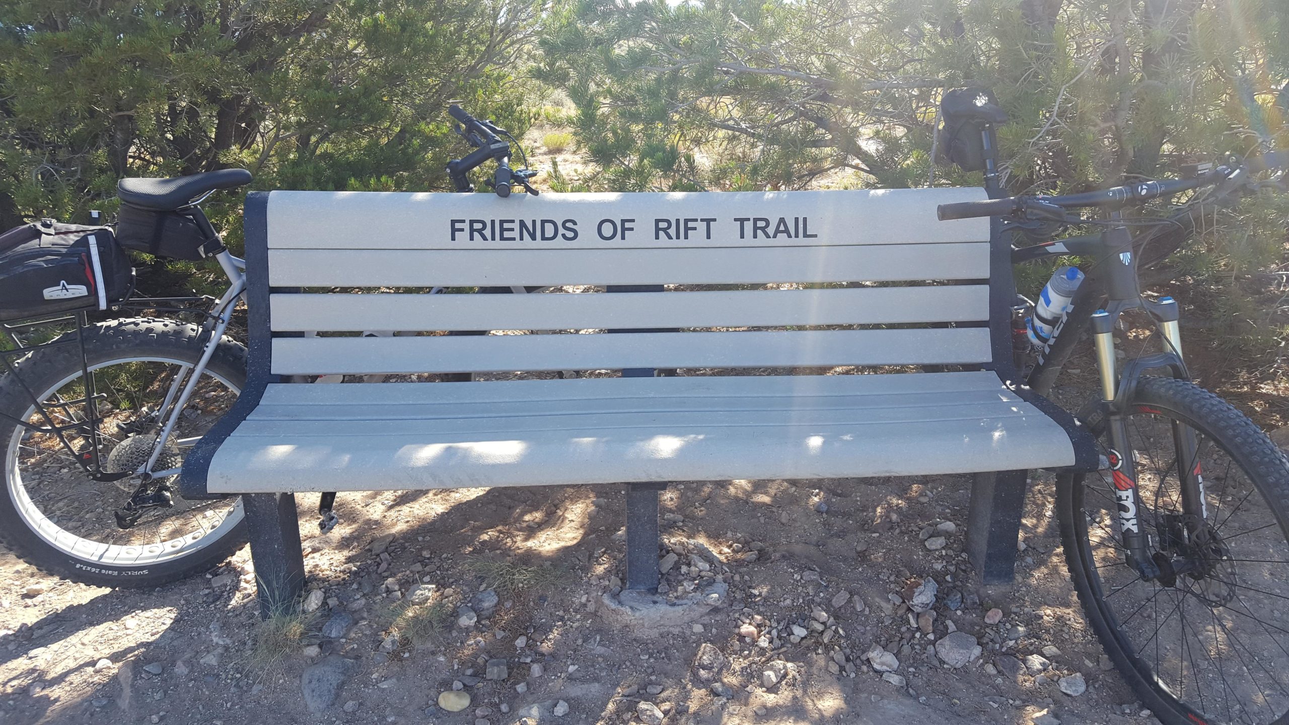 A grey bench with the inscription "FRIENDS OF RIFT TRAIL," surrounded by greenery, with two bicycles parked nearby. The scene captures a sunny day in an outdoor setting, emphasizing a welcoming spot for trail enthusiasts. Taos Valley Overlook mountain bike trail.