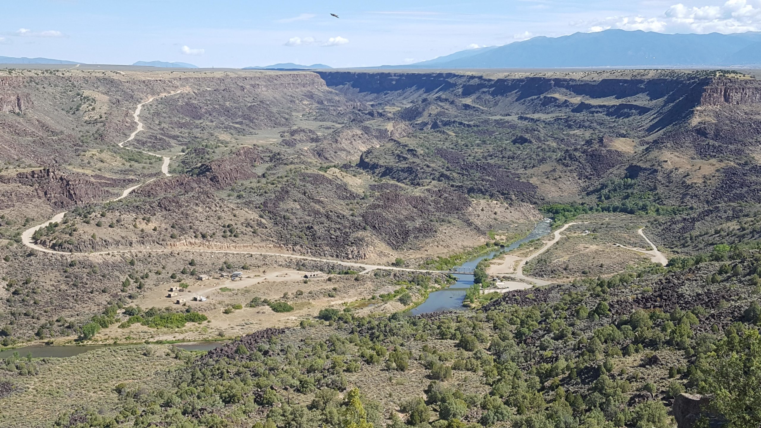 A panoramic view of a rugged canyon landscape with winding dirt roads, lush greenery near a river, and distant mountains under a clear blue sky. The scene captures the natural beauty and varied terrain of the area. Taos Valley Overlook mountain bike trail.
