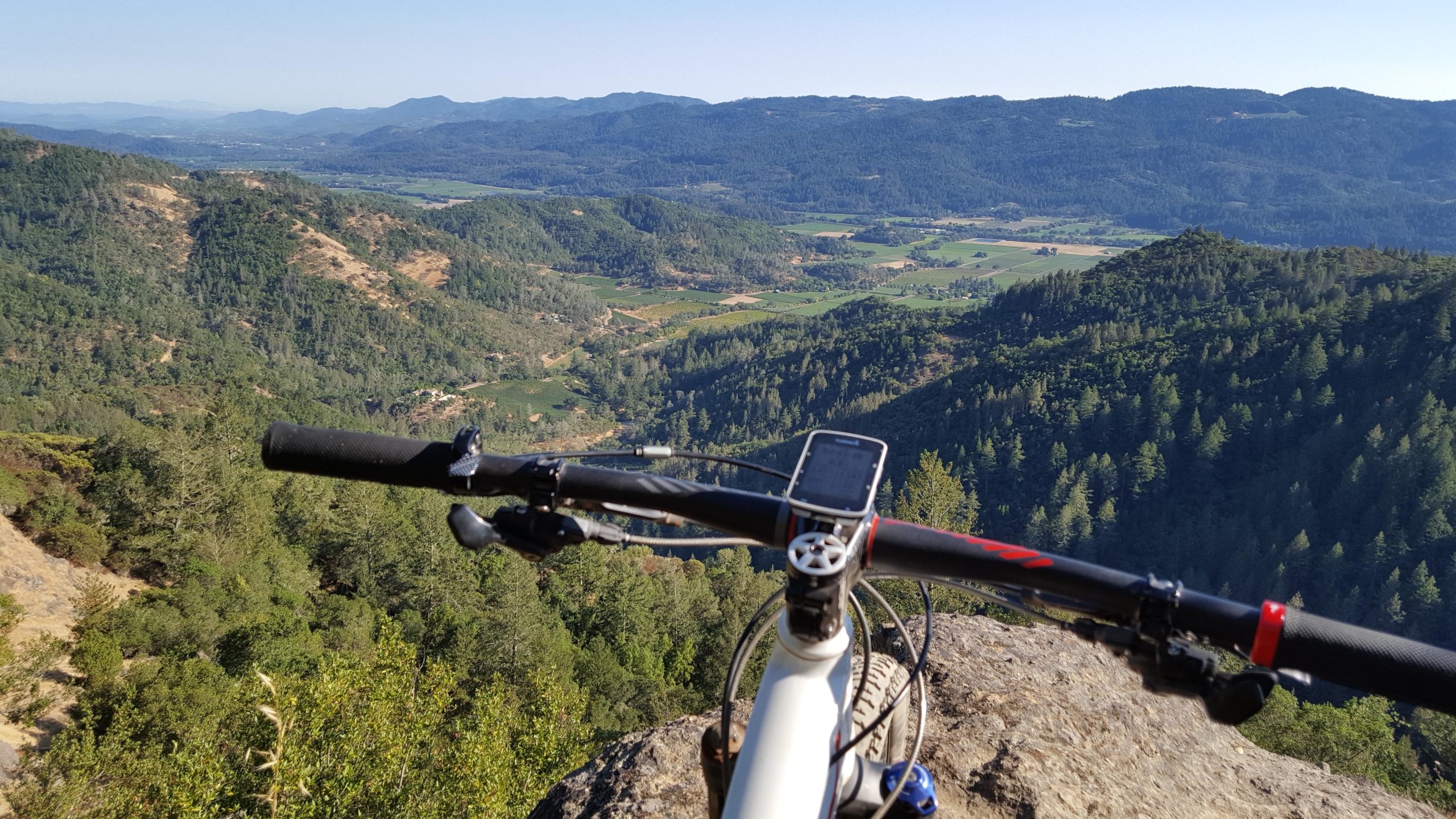 A mountain bike is positioned on a rocky outcrop, overlooking a vast, green valley surrounded by rolling hills and distant mountains under a clear blue sky. The bike's handlebars and GPS device are prominently visible in the foreground, while lush trees and farmland stretch into the horizon. Oat Hill Road mountain bike trail.