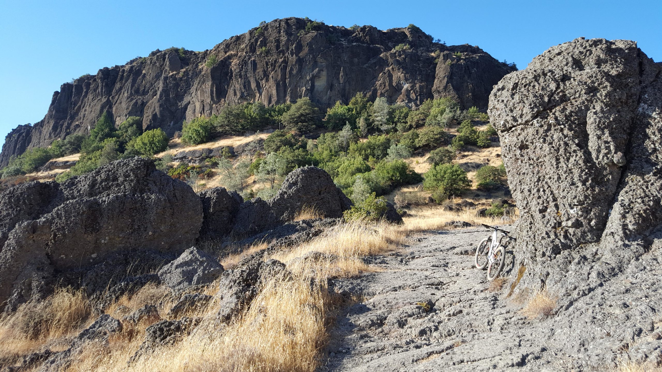 A rocky landscape featuring steep cliffs in the background, with patches of greenery and dry grass. In the foreground, a dirt path is visible, along with a mountain bike leaning against a large rock. The clear blue sky overhead enhances the scene. Oat Hill Road mountain bike trail.