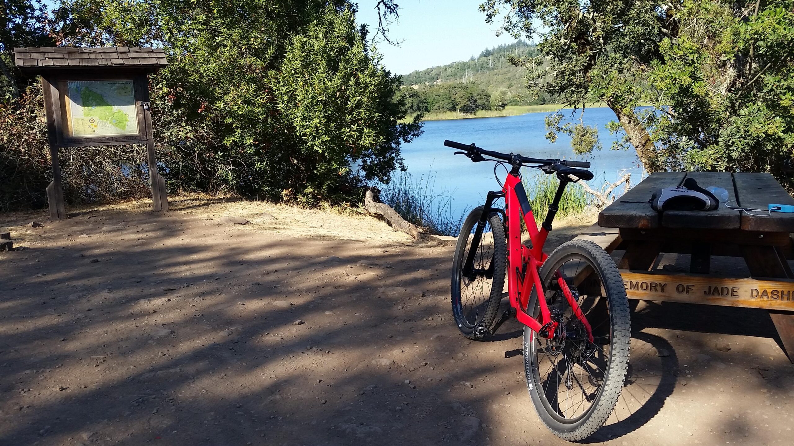 Trek Fuel EX 7 29: A red mountain bike leaning against a wooden picnic table along a dirt path. In the background, a tranquil lake surrounded by lush greenery can be seen, with a map display board nearby. Sunlight filters through the trees, creating a peaceful outdoor setting.