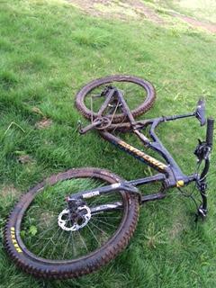 Santa Cruz Bronson C: A black mountain bike lying on its side in a grassy area, with mud on the tires and frame. The front wheel is turned, and the bike appears to have been abandoned or fallen over.