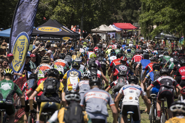 A crowded scene at a mountain bike racing event, featuring numerous participants in colorful jerseys and helmets, riding through a grassy area. Spectators can be seen in the background, with tents and banners promoting the NorCal High School Mountain Bike Racing League. The atmosphere is lively, showcasing the excitement of the competition.