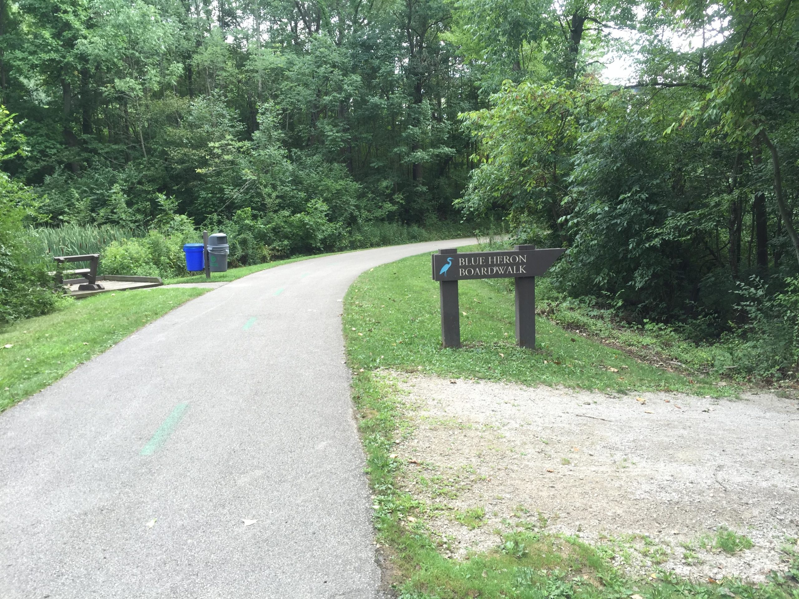 A paved pathway leading into a wooded area, with a sign marking the entrance to the "Blue Heron Boardwalk." There are park benches nearby, along with trash and recycling bins. Lush green trees and shrubs flank the path on either side, creating a serene, natural setting. Ohio And Erie Canal mountain bike trail.