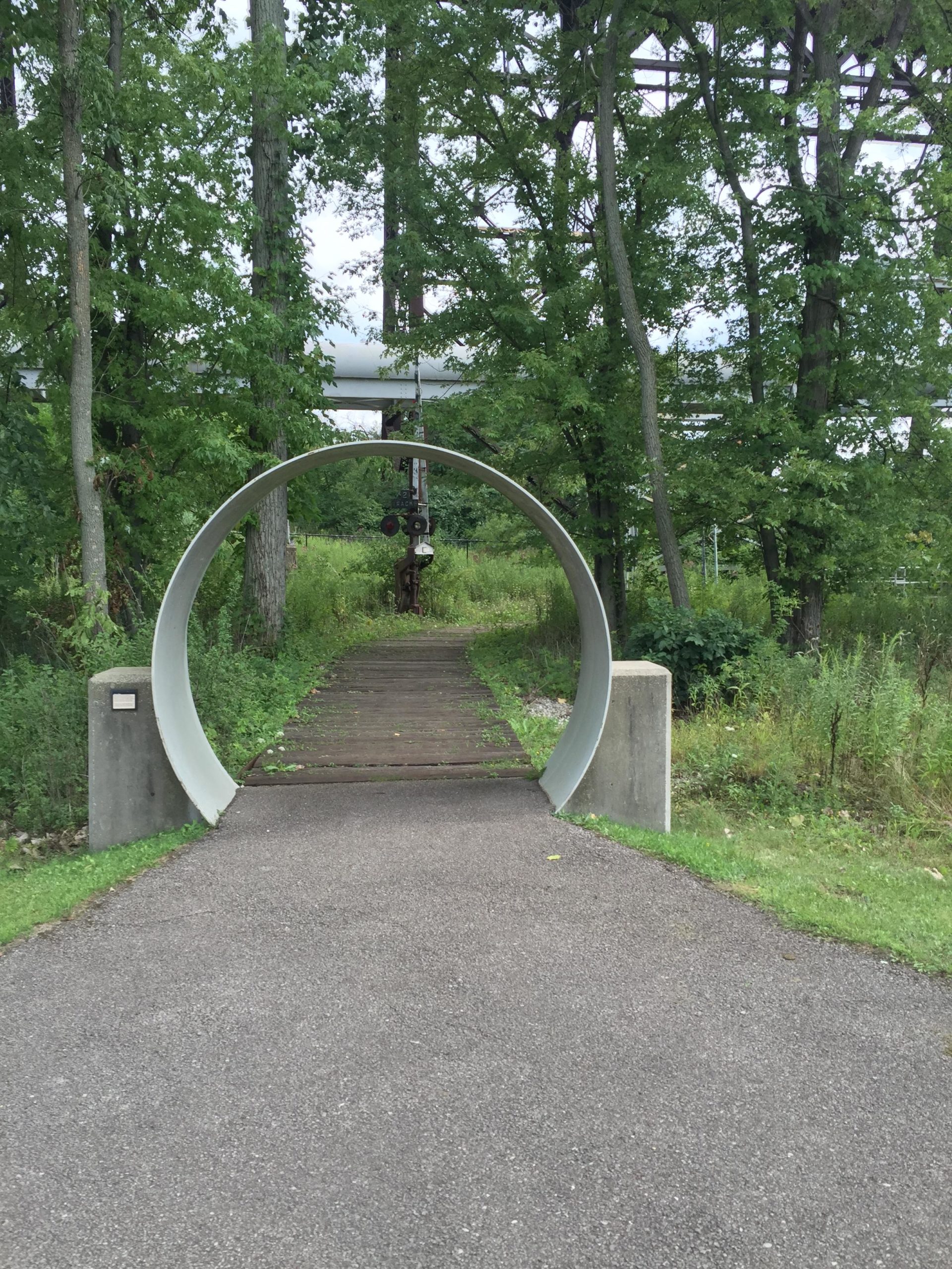 A circular concrete archway stands at the entrance of a pathway surrounded by lush greenery. The trail is bordered by trees and features a mix of gravel and grass, leading into the woods. In the background, a glimpse of a structure can be seen above the trees. Ohio And Erie Canal mountain bike trail.